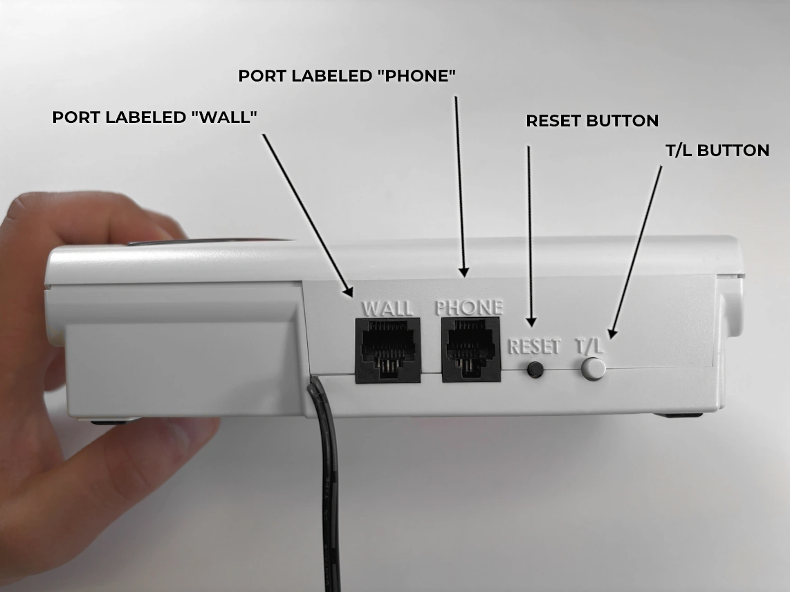holding a base unit in hand with arrows pointing at port labeled 'Wall', port labeled 'PHONE', reset button, and t/l button.